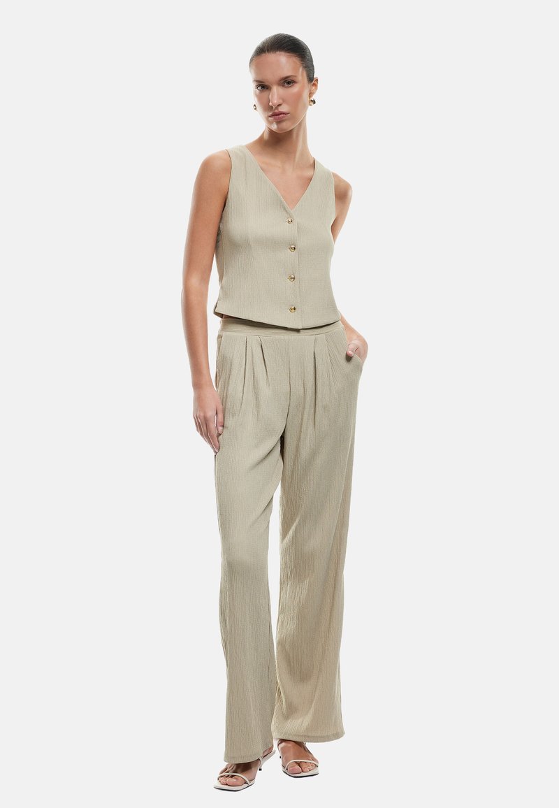 Mujer vistiendo un top sin mangas beige abotonado y pantalones de pierna ancha a juego, de pie con una mano en el bolsillo, fondo neutro.