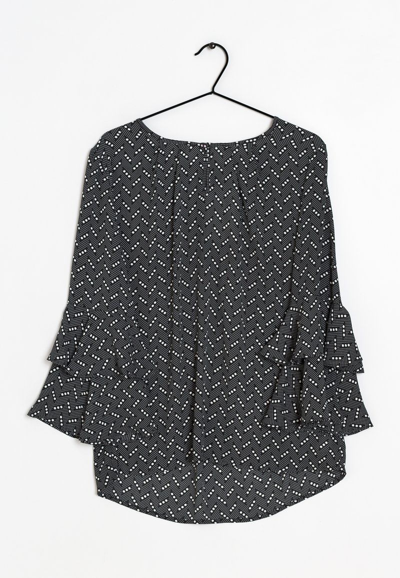 Mustavalkoinen pusero, jossa on kalanruotokuvio, kellohihat ja pyöreä pääntie, ripustettuna mustalle koukulle valkoista taustaa vasten.