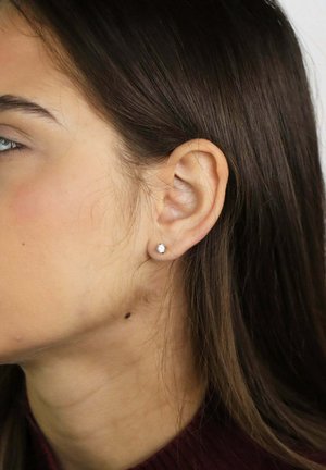 Oreille gauche d'une femme avec une petite boucle d'oreille ronde, cheveux bruns foncés tirés derrière l'oreille, et un grain de beauté visible sur le cou.