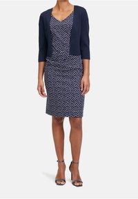 Marinblå prickig klänning med en åtsittande siluett, tillsammans med en kort marinblå cardigan. Har en V-ringning och trekvartslånga ärmar.