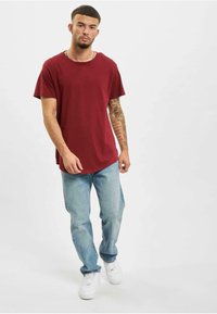 Bordeaux T-shirt med korte ærmer og rund hals; parret med lyseblå jeans og hvide sneakers; modellen står på en neutral baggrund.