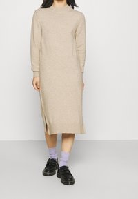 Beżowa dzianinowa sukienka z długimi rękawami i wysokim kołnierzem, z bocznymi rozcięciami. Połączona z czarnymi butami i jasnofioletowymi skarpetkami.