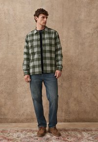 Groene en witte geruite overhemd met knoopsluiting, gecombineerd met blauwe spijkerbroek en bruine schoenen, staand tegen een gestructureerde neutrale achtergrond.