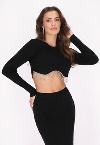 Fekete bordázott crop top, hosszú ujjakkal, hullámos hemvonalra ezüst frufru díszítéssel. Az anyaga puhanak és texturáltak tűnik.