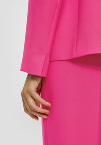 Garnitur z tkaniny w kolorze fuksji o fakturowanym wykończeniu; widoczna ręka dotykająca szwu marynarki, z dopasowanym detalem na rękawie.