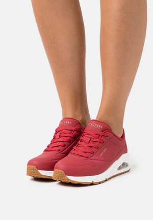Jambes portant des baskets Skechers rouges avec des semelles intermédiaires blanches et des semelles extérieures en caoutchouc teinté, posées sur un fond clair uni.