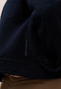 Detaljbild av navyblå texturerad ytterjacka med en svart knapp och en etikett med texten "CECIL OUTERWEAR" över bruna byxor.