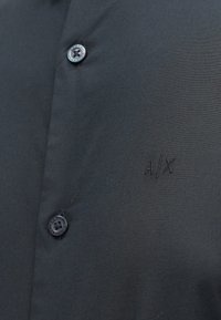 Crna majica s glatkom teksturom, dva gumba i izvezenom oznakom "A/X" s lijeve strane. Moderni i uski dizajn.