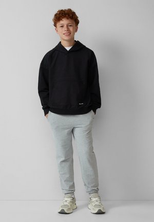 Schwarzer Kapuzenpullover mit einer Fronttasche, kombiniert mit grauen Jogginghosen; steht vor einem schlichten Hintergrund und trägt helle Sneakers.