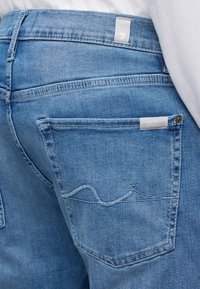 Gros plan sur une personne portant un jean en denim bleu clair avec un motif de vague cousu sur la poche arrière et une petite étiquette en tissu blanc.