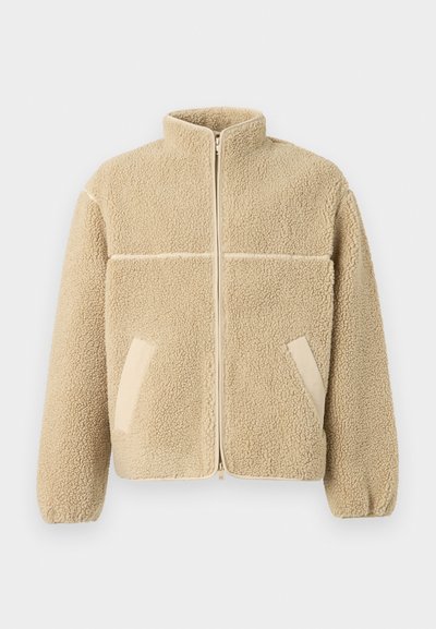 Beige fliisjakker kõrge kaelusega, ees tõmblukuga ja kahe küljetaskuga. Omab tekstuuri, pehmet kangast ja kontrastpaneeli.
