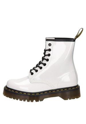 Biały błyszczący but bojowy z czarnymi sznurowadłami, żółtym przeszyciem, grubą brązową podeszwą z rowkami oraz czarno-żółtą pętlą z tyłu.