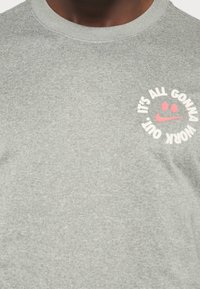 Šedé bavlněné tričko s kulatým výstřihem. Obsahuje grafiku v růžové a bílé, která říká "VŠECHNO SE TO DOBŘE VYŘEŠÍ" s detailním úsměvem.