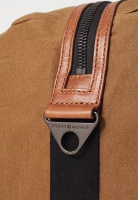 Borsa di tessuto marrone con una cerniera nera, cinturino in pelle beige e un'etichetta metallica incisa con "TOMMY HILFIGER". Texture complessiva liscia.