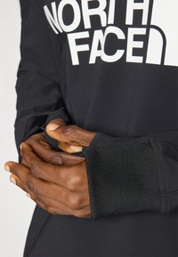 Zwarte longsleeve shirt met opvallend wit "North Face" logo. Heeft een getailleerd ontwerp en ribbelmanchetten met duimgaten. Textuurdetail van de stof is zichtbaar.