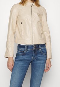 Ljus beige läderjacka med en kortad design, som har en dragkedja fram, två fickor med dragkedja och ett kviltat mönster på axlarna.