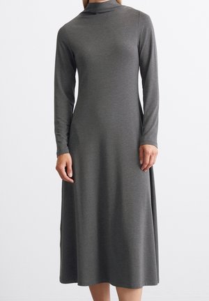 Vrouw draagt een donkergrijze jurk met lange mouwen, hoge hals en enkel lengte, staande tegen een effen witte achtergrond.