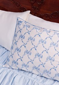 Hvidt silke pudebetræk med blåt sløjfemønster og blå kant, der ligger på lyseblåt lagentæppe med flæser. Blød, glat tekstur.