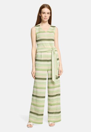 Kvinde stående iført en ærmeløs grøn mønstret jumpsuit med vide ben og et matchende bælte bundet om taljen, kombineret med hvide sandaler.