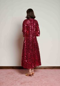 Γυναίκα με καστανά μαλλιά, φοράει μακρύ κόκκινο φόρεμα με παγιέτες και ταμπά ψηλοτάκουνα παπούτσια, στέκεται μπροστά σε έναν λευκό τοίχο πάνω σε ροζ χαλί.