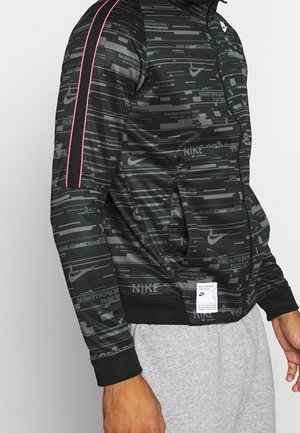 Black Nike sports jacket with all-over logo pattern, pink stripes on sleeves, front pockets, and a white label near the waist, worn with gray pants.