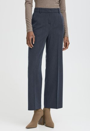 Pantalon large gris foncé porté avec un haut beige à manches longues et des bottines pointues à talons hauts couleur fauve sur un fond uni.