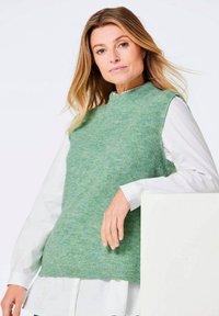 Ärmelloser Strickpullover in Hellgrün mit einem strukturierten Rippenmuster, über einem weißen Hemd mit Kragen getragen, das ein hochgeschlossenes Design aufweist.