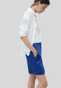 Biela ľanová košeľa s gombíkmi, nosená otvorená cez priliehavý biely top a modrú ľanovú sukňu. Sukňa má bočné vrecká a rovný strih.