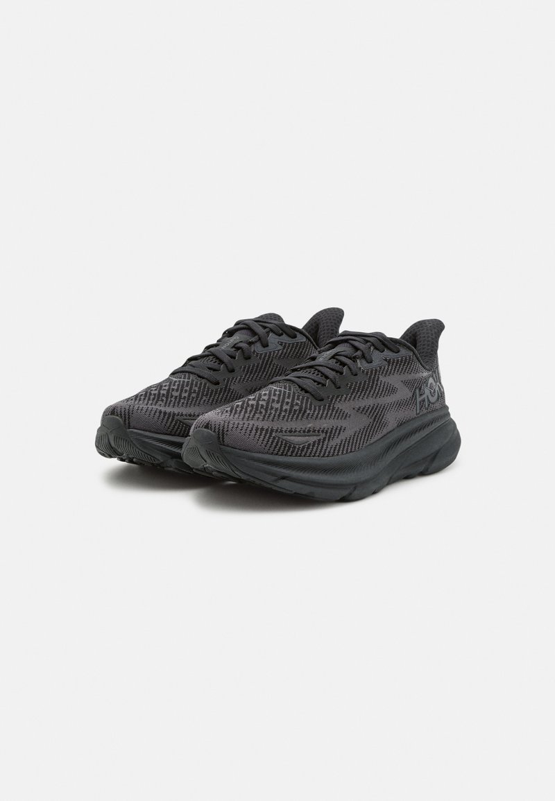 Schwarze Sportschuhe mit einem strukturierten Mesh-Obermaterial, einer robusten Gummisohle und flachen Schnürsenkeln. Verfügt über ein dezentes Logo an der Seite und ein gemustertes Design.