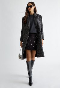 Cappotto grigio testurizzato, maglione scuro, gonna mini nera con decorazioni, stivali lucidi fino al ginocchio e borsa a tracolla argentata. Occhiali da sole inclusi.