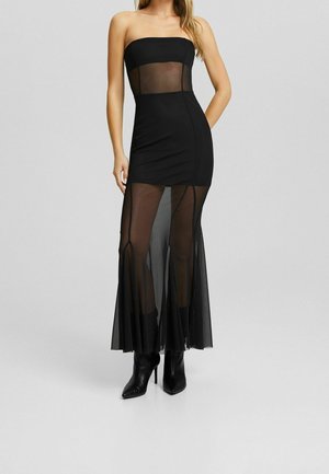 Robe noire sans bretelles avec corsage en maille transparente et sur-jupe longue, portée avec des bottines noires à talons sur un fond clair.