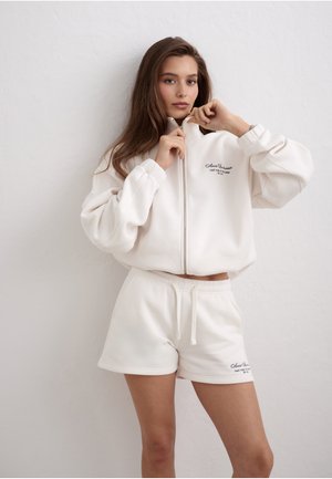 Młoda kobieta z długimi brązowymi włosami zapinająca biały rozpinany na zamek sweatshirt, ubrana w białe szorty ze sznurkiem, stojąca na tle gładkiej ściany.