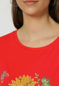Camiseta de algodón roja con cuello redondo, decorada con flores amarillas bordadas y una mariposa en hilo colorido. Textura suave.