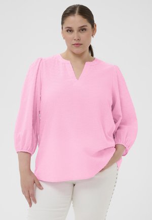 Femeie cu păr castaniu prins într-o coadă joasă, purtând o bluză roz texturată cu mâneci 3/4 și pantaloni albi pe un fundal simplu.