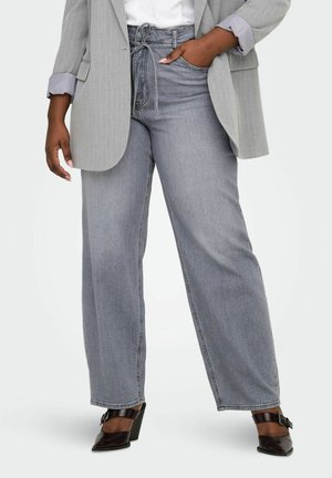 Kvinne iført grå vidbente jeans med høyt liv, grå pinstripeblazer, hvit skjorte og svarte pumps, med hånden i lommen.