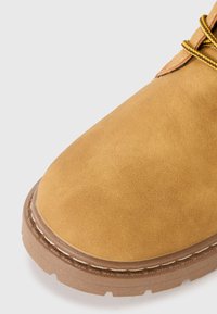 Bota de ante marrón claro con punta redonda, suela de goma marrón claro contrastante y detalles cosidos a lo largo del borde. Los cordones son amarillos y marrones.
