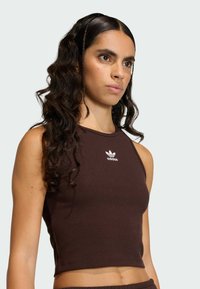 Hnedý rebrovaný crop top bez rukávov s okrúhlym výstrihom. Na hrudi je biely nápis Adidas. Hladký materiál.
