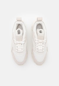 Valkoiset ja vaaleanbeigekengät urheiluun, joissa on nauhat, verkkomateriaalia ja mokkasiiniedustuksia, kuvattuna ylhäältä valkoista taustaa vasten.