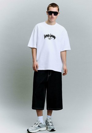 Giovane uomo con indosso una T-shirt bianca oversize con grafica, pantaloncini neri a gamba larga, calzini bianchi, sneakers e grandi occhiali da sole neri, in piedi davanti a uno sfondo grigio.
