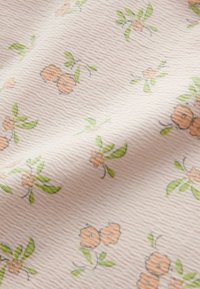 Blødt tekstureret stof med små orange blomster og grønne blade spredt ud over en lys beige baggrund.