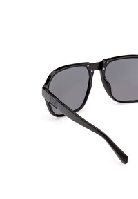 Lunettes de soleil à monture carrée noire avec des verres gris foncé et des branches épaisses, ornées d'un petit logo en métal sur la branche.