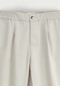 Pantalon beige clair avec une taille élastique, fermeture avant à bouton métallique, et braguette cachée, fabriqué en tissu lisse.