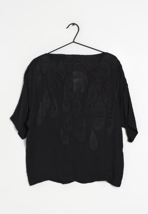 Camisa negra de silueta holgada con un escote amplio, mangas cortas y patrones texturizados que parecen gotas. Cuelga de un gancho negro.