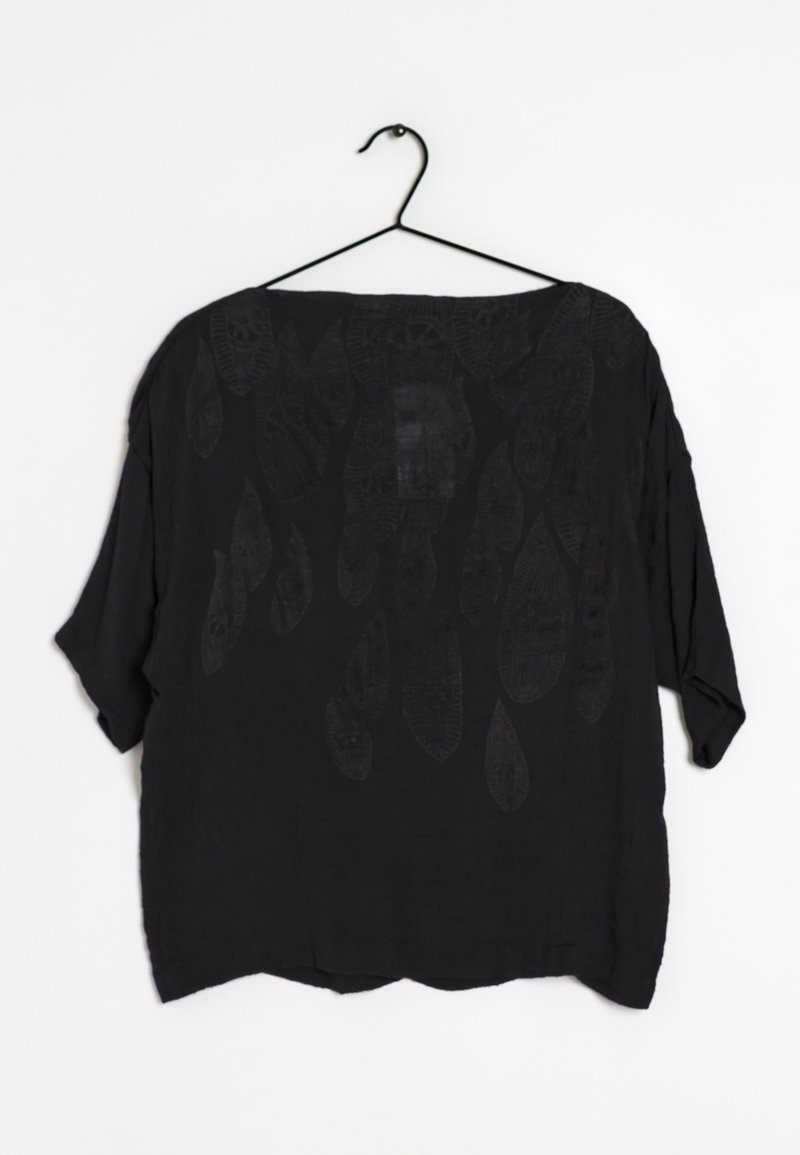 Chemise noire ample avec un large décolleté, des manches courtes et des motifs texturés ressemblant à des gouttes. Suspendue à un crochet noir.