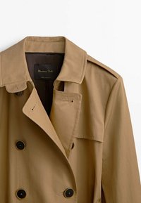 Tan trench coat with a double-breasted design, large black buttons, wide lapels, and shoulder epaulets, made from smooth fabric.