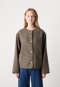 Brun hørnbåndsjakke med rund hals, store frontlommer, sølvknapper og vide ærmer, parret med blå denim jeans.