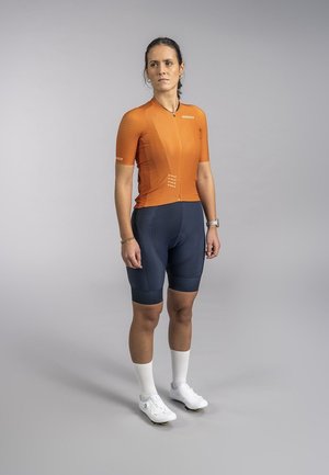 Oranje fietsshirt met korte mouwen en een ritssluiting aan de voorkant, gecombineerd met marineblauwe fietsshorts en witte fietsschoenen. Glad materiaal met een sportieve pasvorm.