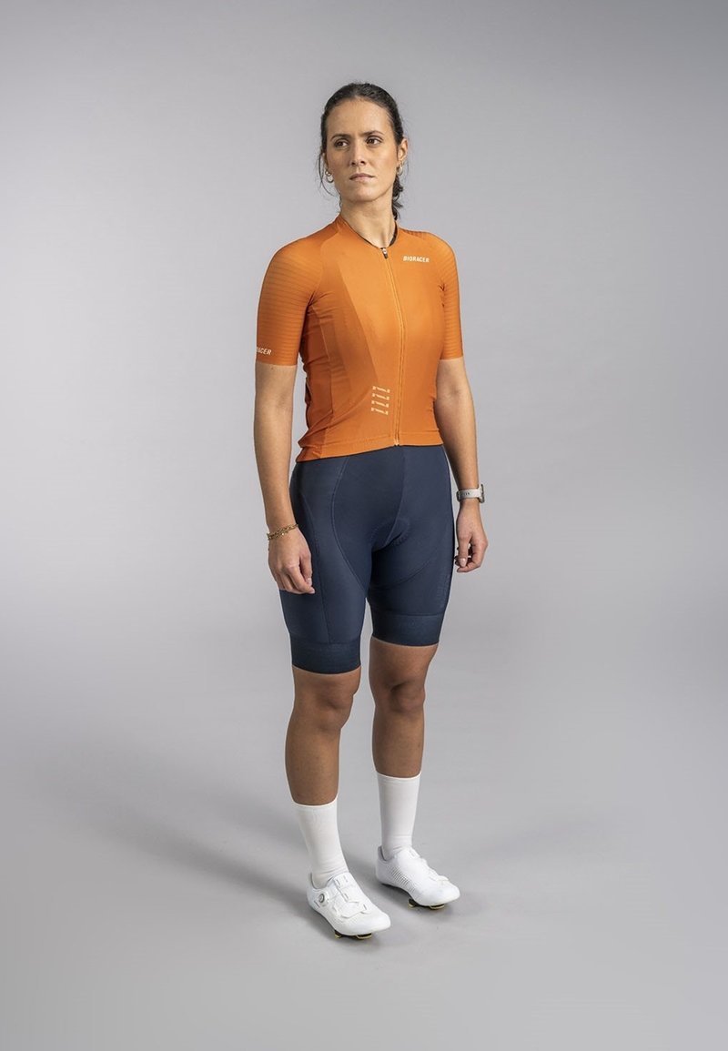 Oranje fietsshirt met korte mouwen en een ritssluiting aan de voorkant, gecombineerd met marineblauwe fietsshorts en witte fietsschoenen. Glad materiaal met een sportieve pasvorm.
