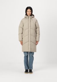 Casaco puffer bege com capuz, apresentando um padrão acolchoado e fecho de correr frontal, combinado com jeans azuis e sapatos pretos. Textura de tecido suave.