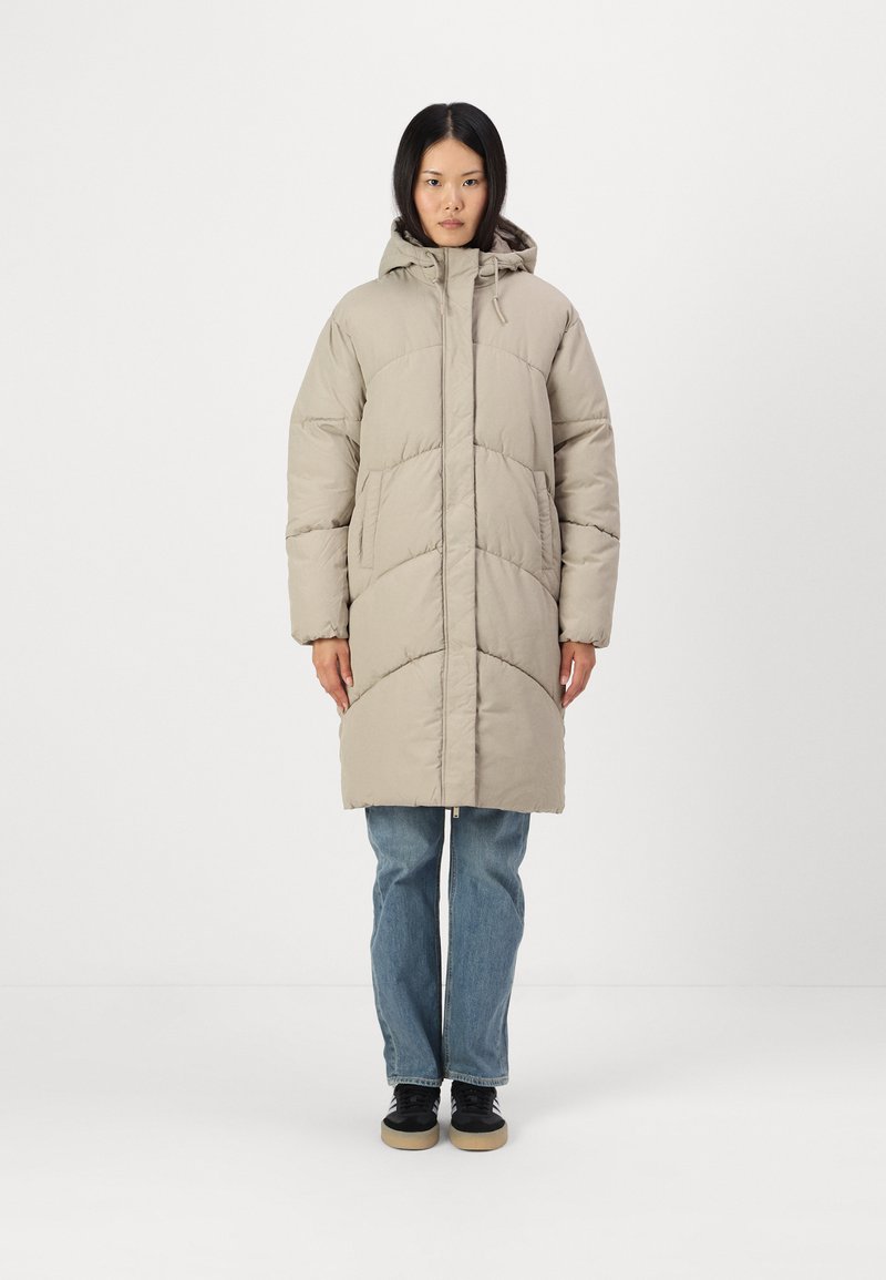 Casaco puffer bege com capuz, apresentando um padrão acolchoado e fecho de correr frontal, combinado com jeans azuis e sapatos pretos. Textura de tecido suave.